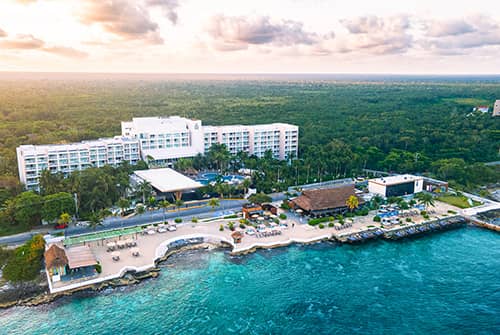 Aerial view of a beachfront resort in Mexico with white buildings, pools, palm trees, and lounge areas along the turquoise shoreline—perfect for meetings and conference events amid lush greenery under a partly cloudy sky.