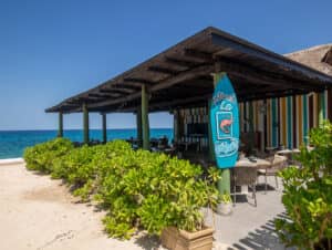 Un restaurante junto a la playa, con techo de madera y paredes a rayas de colores, se ubica entre exuberantes arbustos verdes sobre arena, con vistas a un océano azul brillante cerca de los mejores resorts de México; ideal para reuniones informales o eventos de conferencias. Un letrero con forma de tabla de surf anuncia el Restaurante La Calypso.