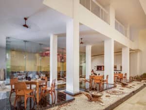 Interior de restaurante moderno con techos altos, columnas blancas y mamparas de cristal, perfecto para conferencias y reuniones. Mesas de madera sobre plataformas oscuras rodeadas de piedras blancas crean un ambiente minimalista y abierto, ideal para hoteles y resorts en México.