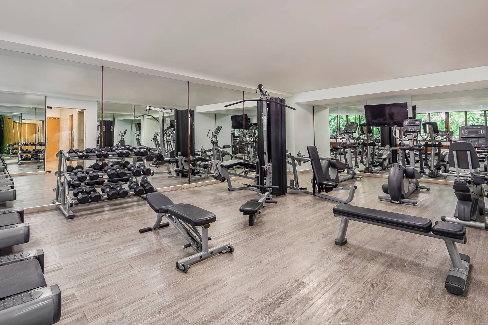 A spacious gym in Mexico resorts with wooden flooring, mirrors, free weights, benches, resistance machines, treadmills, ellipticals, and large windows letting in natural light—perfect for guests attending conferences or meetings.