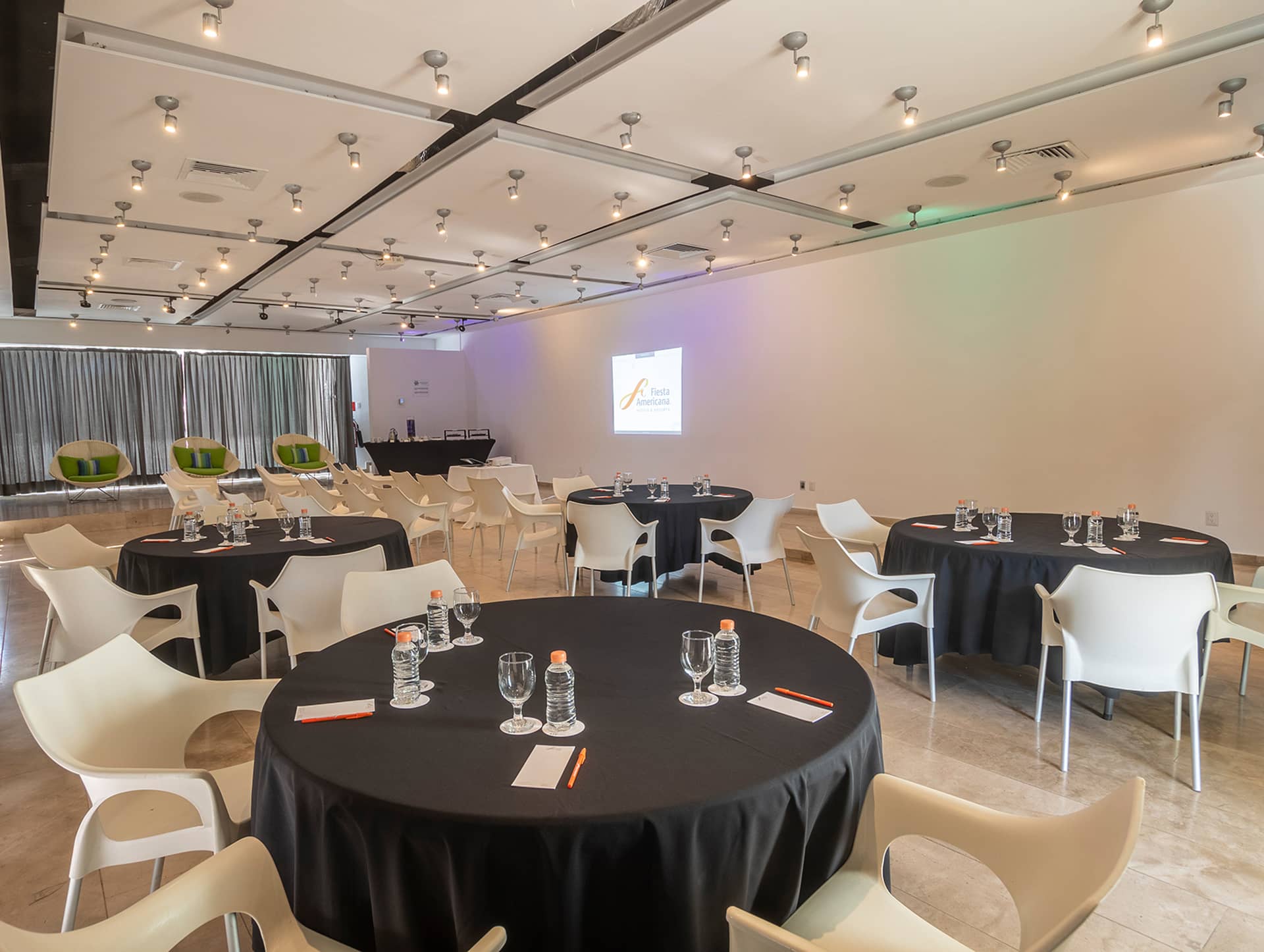 A modern conference room at Mexico resorts, with round tables covered in black cloths, set with glasses, water bottles, and notepads. White chairs surround the tables, perfect for meetings or conference events, with a projection screen on a white wall.