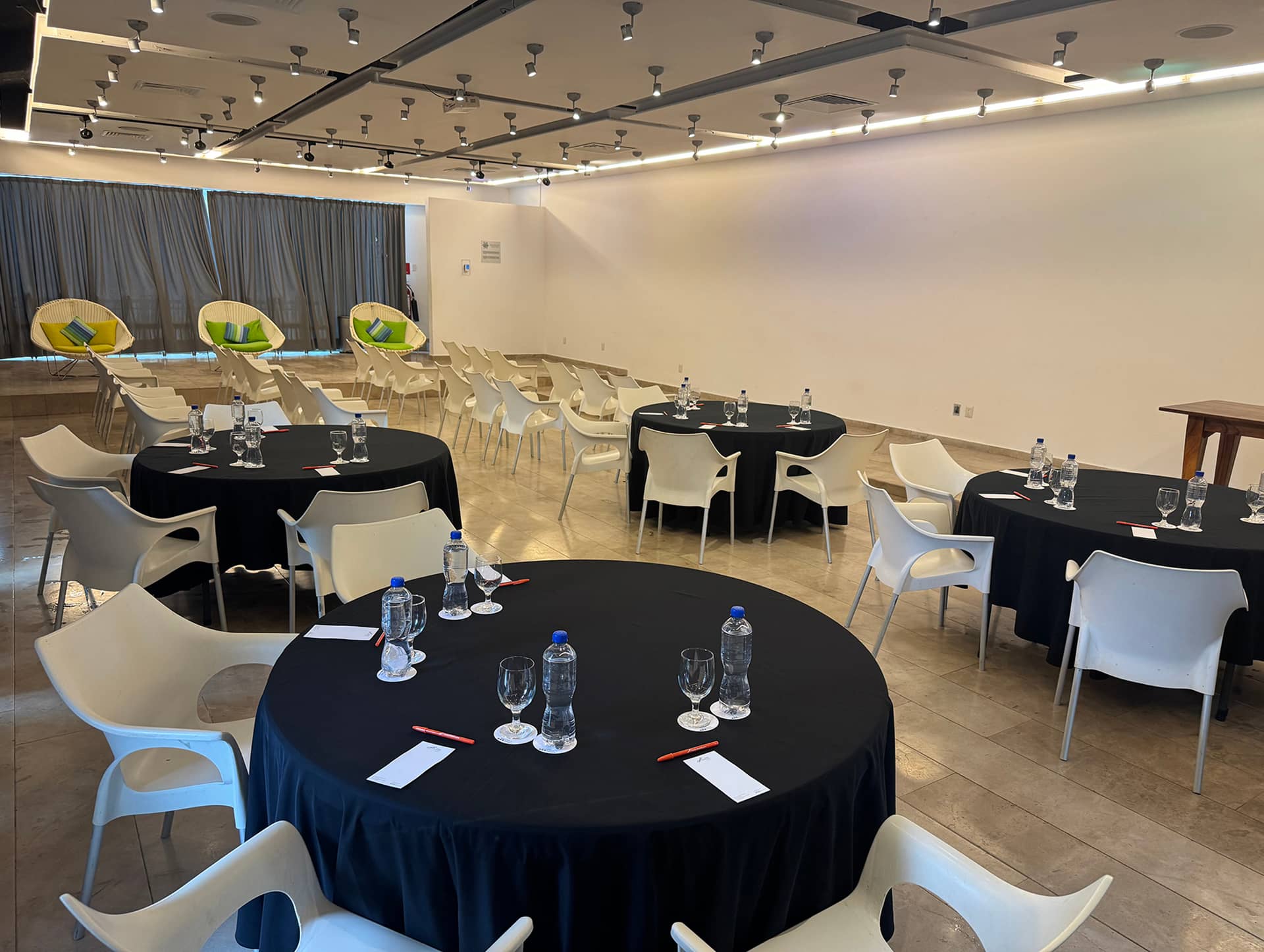 A conference room at a Mexico resort with round tables covered in black cloths, set for meetings with water bottles, glasses, and notepads. White chairs surround the tables, with extra chairs and beanbags at the back of the room.