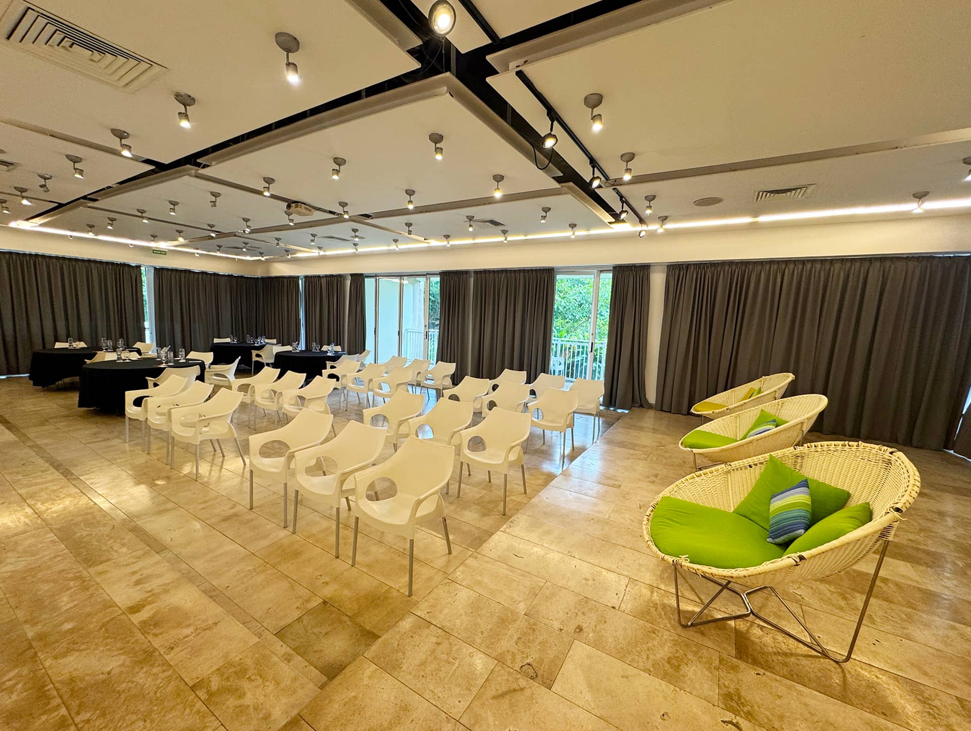 A spacious conference room ideal for meetings at Mexico resorts, featuring rows of white chairs facing a table on a tiled floor. Two round lounge chairs with green cushions sit in the corner, and large windows with closed curtains line the wall.