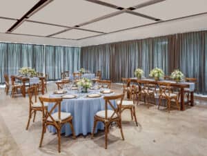 Un salón de banquetes ideal para reuniones o conferencias, con mesas redondas y rectangulares con manteles azul claro, sillas de madera con cojines blancos, centros de mesa florales, grandes ventanales con cortinas grises y suelo de baldosas claras.