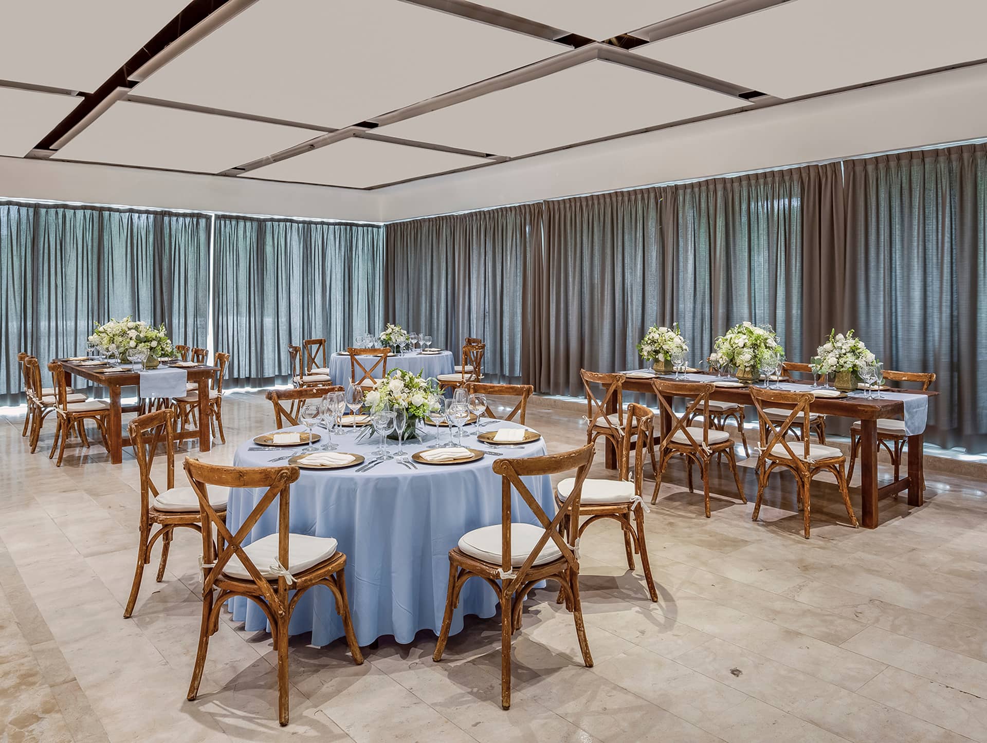 A banquet room ideal for meetings or conference events, featuring round and rectangular tables with light blue tablecloths, wooden chairs with white cushions, floral centerpieces, large windows with gray curtains, and a light tile floor.
