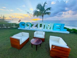 Cuatro sillas de madera con cojines blancos rodean una pequeña mesa redonda sobre el césped, frente a las grandes letras COZUMEL, una palmera y el océano al atardecer: un lugar impresionante para reuniones informales o eventos de conferencias en los mejores complejos turísticos de México.