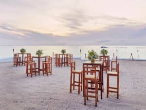 Mesas altas de madera y sillas se disponen en una playa de arena al atardecer para eventos de conferencias, cada mesa decorada con un centro de mesa floral. El océano y un barco a lo lejos se divisan bajo un cielo de tonos pastel, ofreciendo el telón de fondo perfecto para reuniones memorables en México.