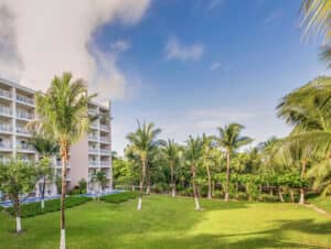 Un frondoso césped verde con palmeras se extiende junto a un hotel mexicano de varias plantas con balcones, perfecto para reuniones y eventos bajo un cielo azul con nubes dispersas.
