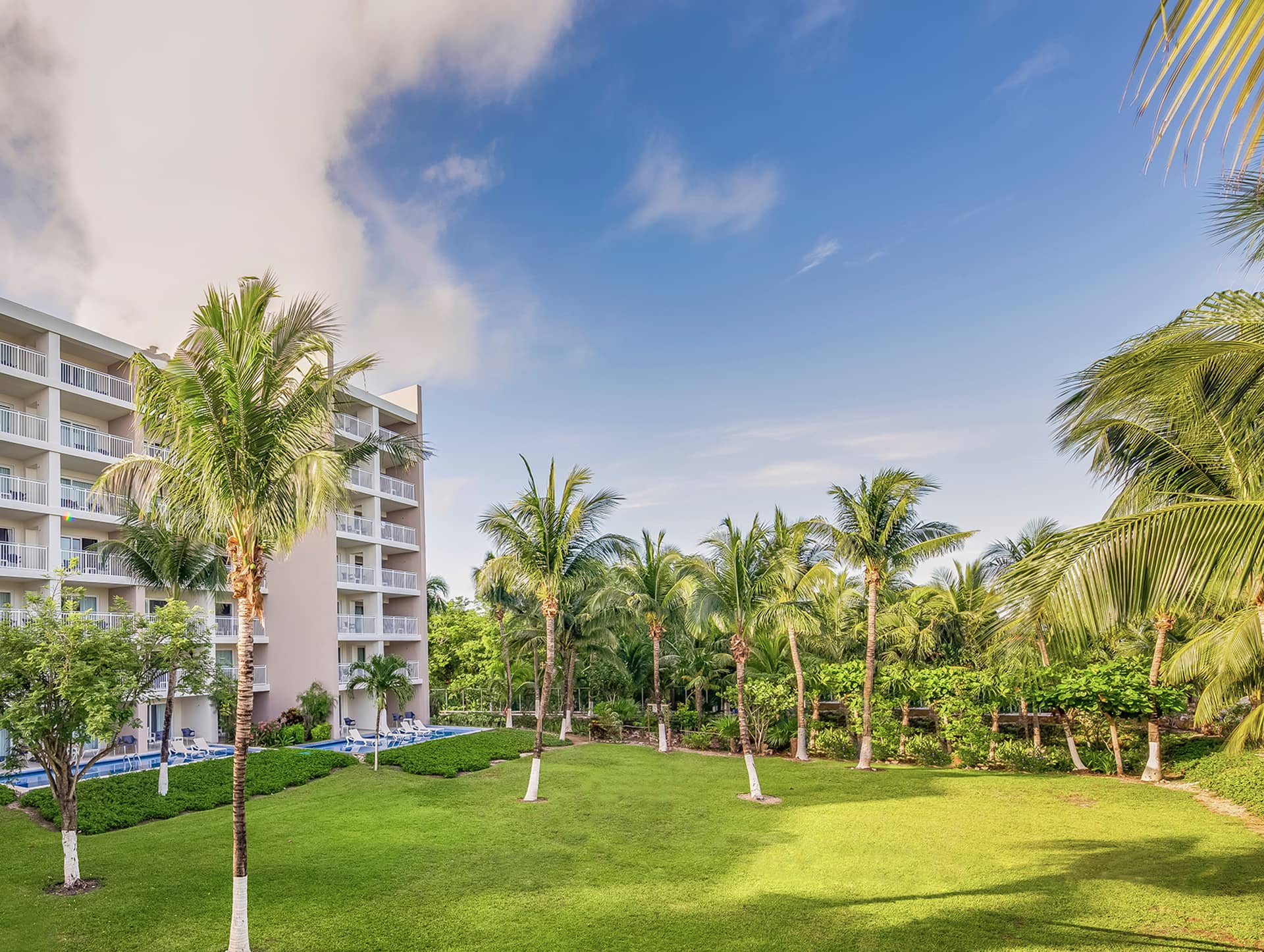 A lush green lawn with palm trees sits beside a multi-story Mexico hotel with balconies, perfect for meetings and conference events under a blue sky with scattered clouds.