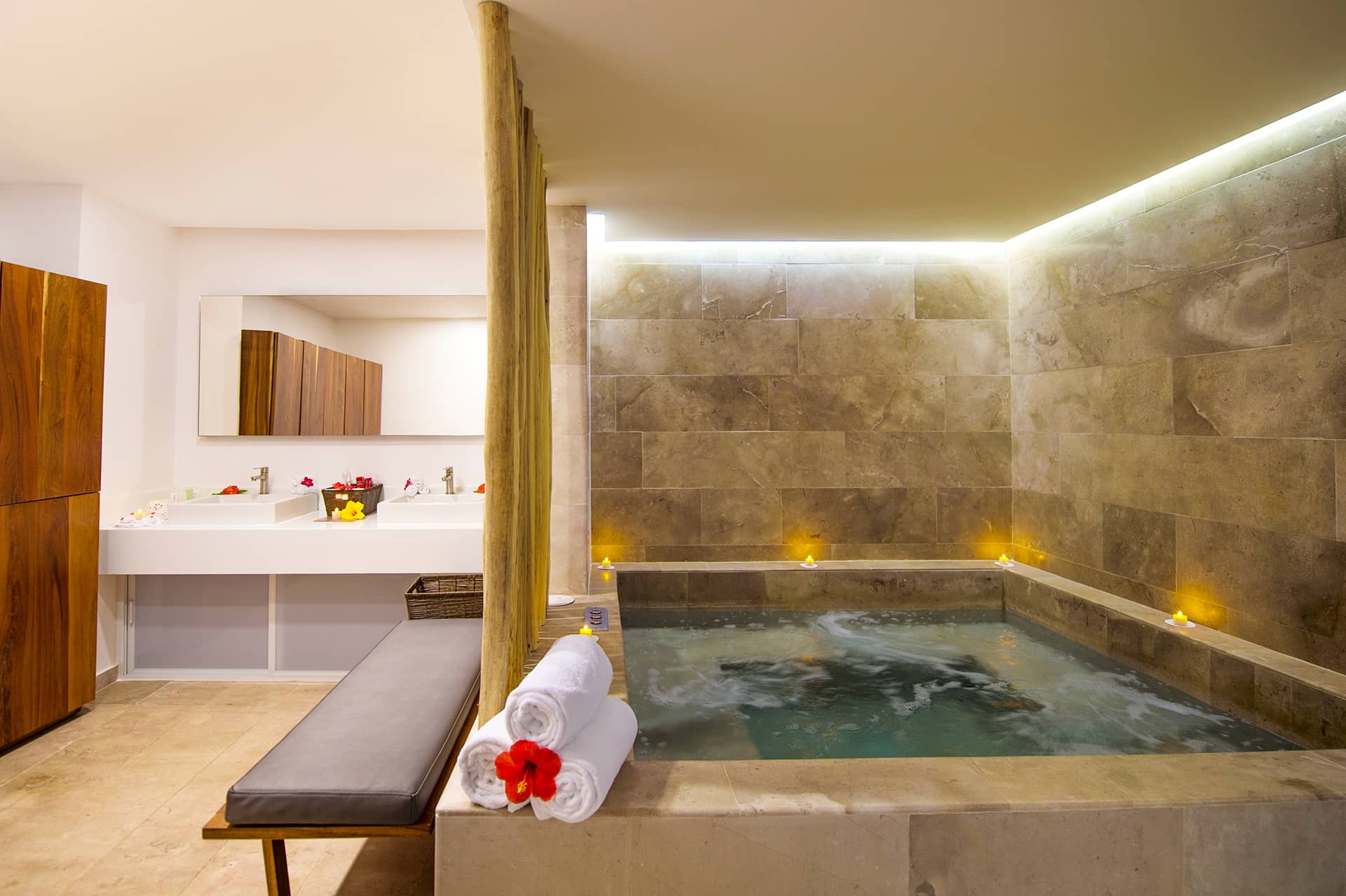 A luxurious spa room in one of Mexico's top resorts, featuring a square stone hot tub with bubbling water, surrounded by lit candles—perfect for relaxing after conference events or meetings. A modern double sink vanity completes the serene setting.