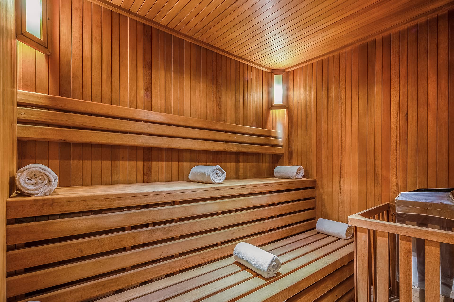 A wooden sauna room with two-tiered benches, three rolled white towels, soft lighting on the walls, and a metal sauna heater in the corner—perfect for relaxing after conference events at Mexico hotels or resorts.