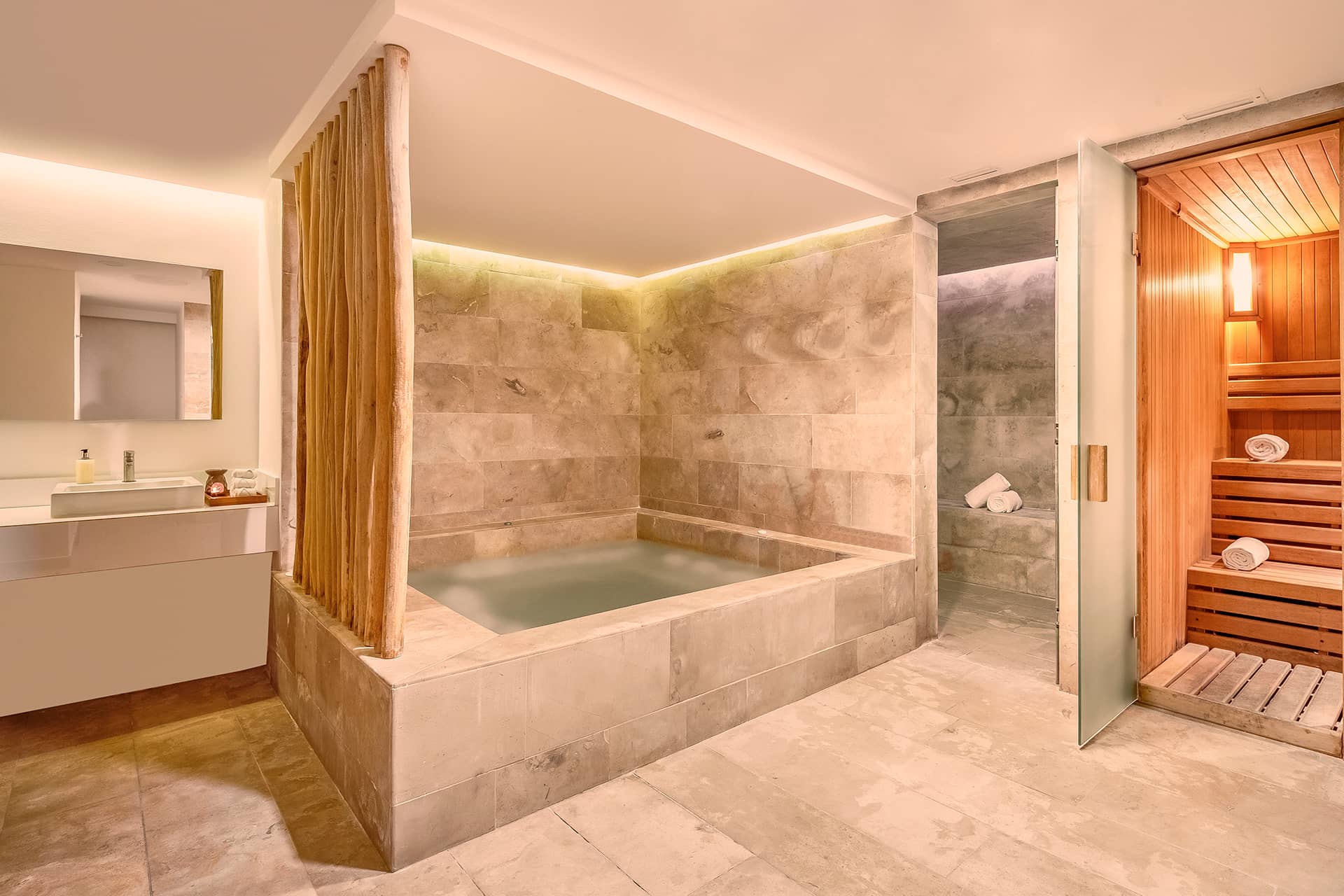 Modern spa room at Mexico resorts with a large stone soaking tub, a vanity with sink and mirror, and a wooden sauna with towels on shelves. The space features warm lighting and neutral tones—perfect relaxation after meetings or conference events.
