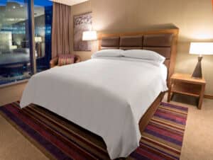 Habitación de hotel moderna con una cama doble bien hecha, cabecero de madera, dos mesitas de noche con lámparas y un sillón, ideal para hoteles en México que organizan reuniones o conferencias, con una gran ventana con vistas a los edificios de la ciudad al atardecer.
