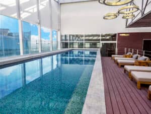 Piscina cubierta de agua cristalina, tumbonas y terraza de madera. Los amplios ventanales ofrecen vistas a la ciudad y luz natural, ideal para relajarse entre reuniones en los mejores hoteles y resorts de México que albergan eventos y conferencias. Modernas lámparas cuelgan del techo.
