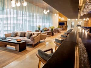 Salón moderno con sofás y sillones beige, mesas de madera y una elegante barra con taburetes altos, ideal para reuniones o eventos. Grandes ventanales con cortinas translúcidas dejan entrar la luz natural, aportando calidez a este espacio de estilo resort.