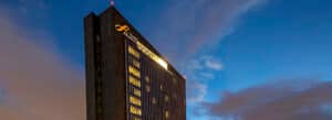Un moderno rascacielos con el logotipo de Budget Insurance se ilumina contra un cielo vespertino azul con nubes dispersas. Varias ventanas están iluminadas, lo que sugiere que se están celebrando reuniones o conferencias en su interior.