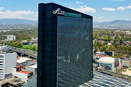 A tall, modern glass skyscraper bearing the Fiesta Americana logo rises above smaller buildings, showcasing one of Mexico’s premier hotels ideal for meetings and conference events, with a cityscape and mountains in the background under a partly cloudy sky.