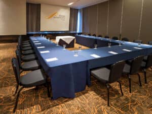 La sala de conferencias del hotel Fiesta Americana en México cuenta con mesas en forma de U cubiertas con manteles azules, preparadas para reuniones con documentos y vasos a mano. Sillas negras y una pantalla de proyección dan la bienvenida a los huéspedes a eventos de conferencias inolvidables en este resort de lujo.