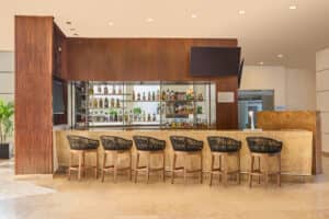 Un moderno bar de hotel con una barra de mármol claro, cinco taburetes de madera y color negro, un gran panel de madera, botellas de licor en estantes de cristal y dos televisores de pantalla plana: perfecto para relajarse después de eventos o reuniones en hoteles y complejos turísticos de México.