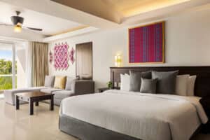 Una espaciosa habitación de hotel en uno de los mejores resorts de México, con una cama grande, sofá gris, mesa de madera y coloridas obras de arte en las paredes. Disfrute de una decoración moderna y pisos de baldosas claras, perfectos para relajarse después de eventos o reuniones.