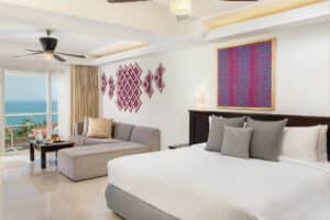 Habitación de hotel moderna con una cama grande, sofá gris y mesa de centro junto a puertas corredizas de cristal que dan a un balcón con vista al mar. Ideal para reuniones o eventos, este resort en México cuenta con coloridas obras de arte geométricas en las paredes blancas.