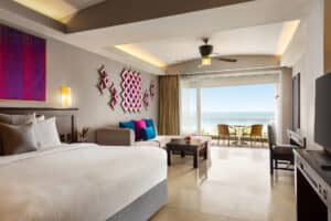 A modern hotel room with a large bed, sofa, and coffee table. Colorful wall art and cushions add accents. Sliding glass doors open to a balcony with chairs and an ocean view—perfect for relaxing after conference events at Mexico resorts.