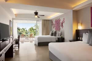 A modern Mexico hotel room with two large beds, a flat-screen TV, desk, and a balcony with chairs overlooking palm trees and greenery—perfect for relaxing after meetings or conference events. The space features neutral tones and decorative wall art above the beds.