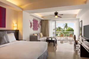 A modern hotel room with a large bed, gray sofa, TV, and wall art. Ideal for conference events or meetings, glass doors open to a balcony with table, chairs, and views of palm trees—perfect for relaxing between sessions at Mexico resorts hotels.