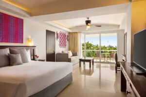 Una moderna habitación de hotel en México con una cama grande, sofá, mesa y televisor. La luminosa decoración incluye coloridos murales, suelo de baldosas y puertas corredizas de cristal que dan a un balcón, ideal para relajarse después de conferencias o reuniones en el resort.