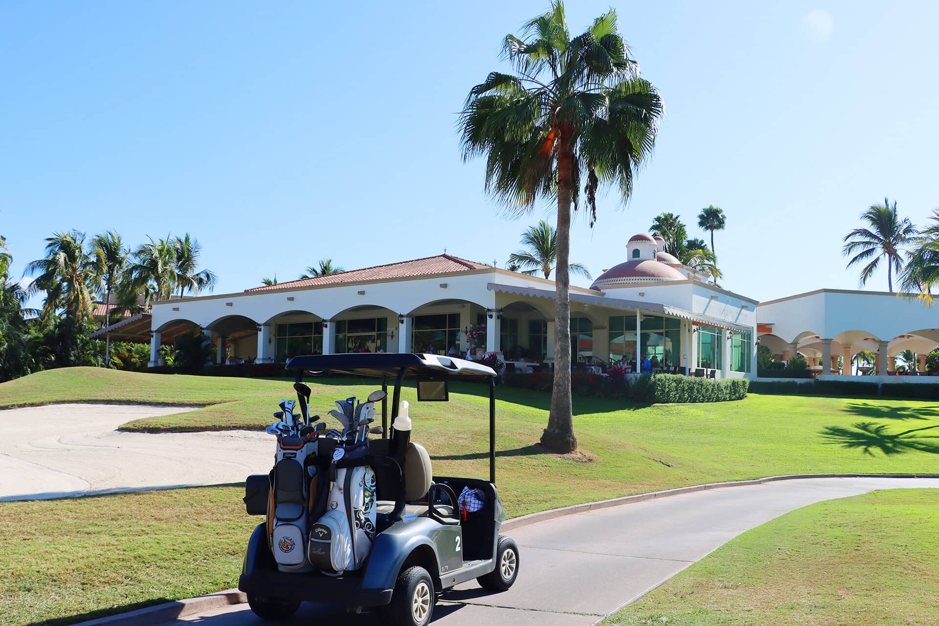 A golf cart with clubs is parked on a paved path near a sand bunker, in front of a clubhouse with arches and palm trees—perfect for relaxing after meetings at one of Mexico's premier conference events resorts.