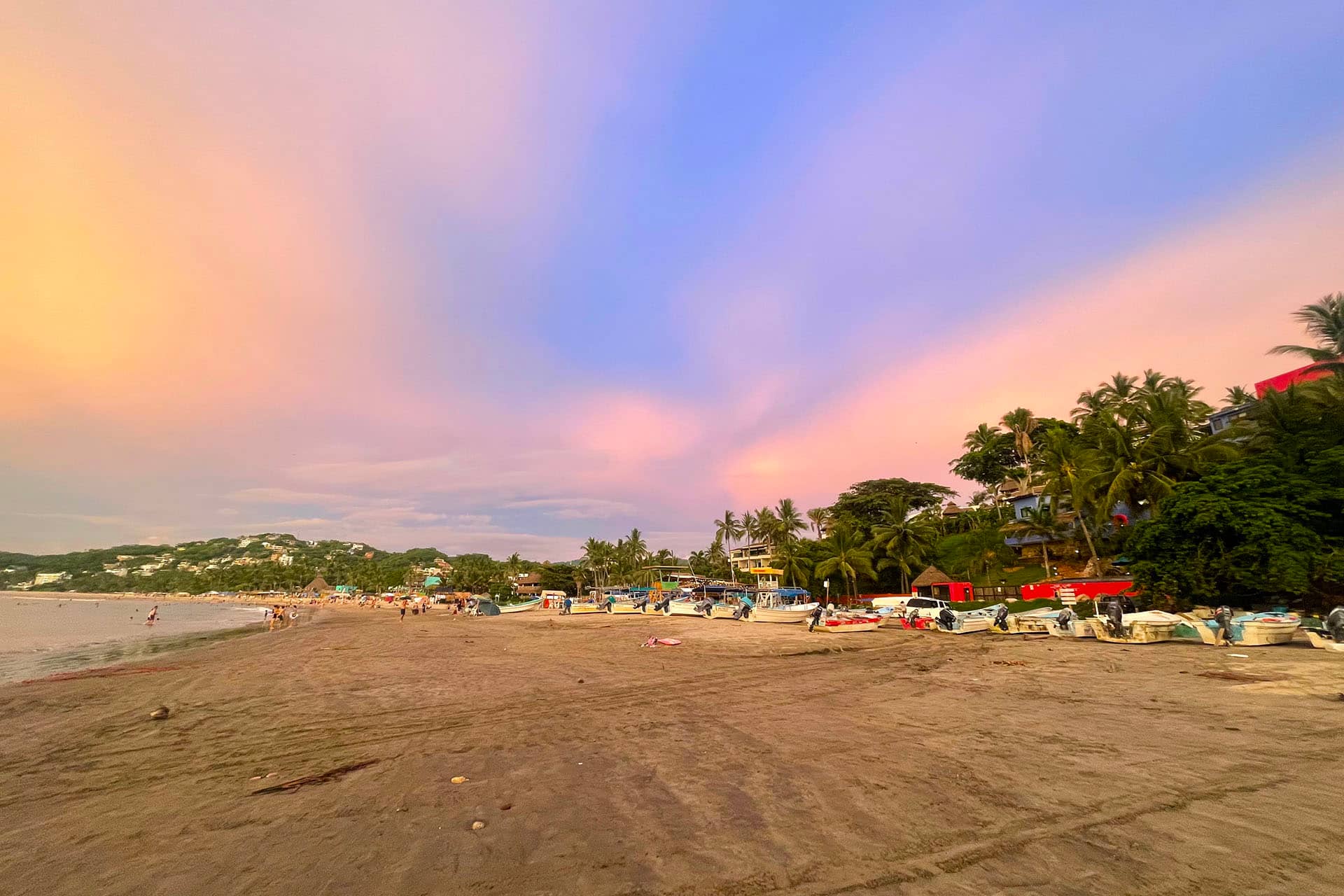Boats line a sandy beach at sunset in Mexico, with palm trees and lush greenery behind them. The softly colored sky overlooks distant hills—an ideal backdrop for unforgettable conference events or relaxing stays at nearby resorts.