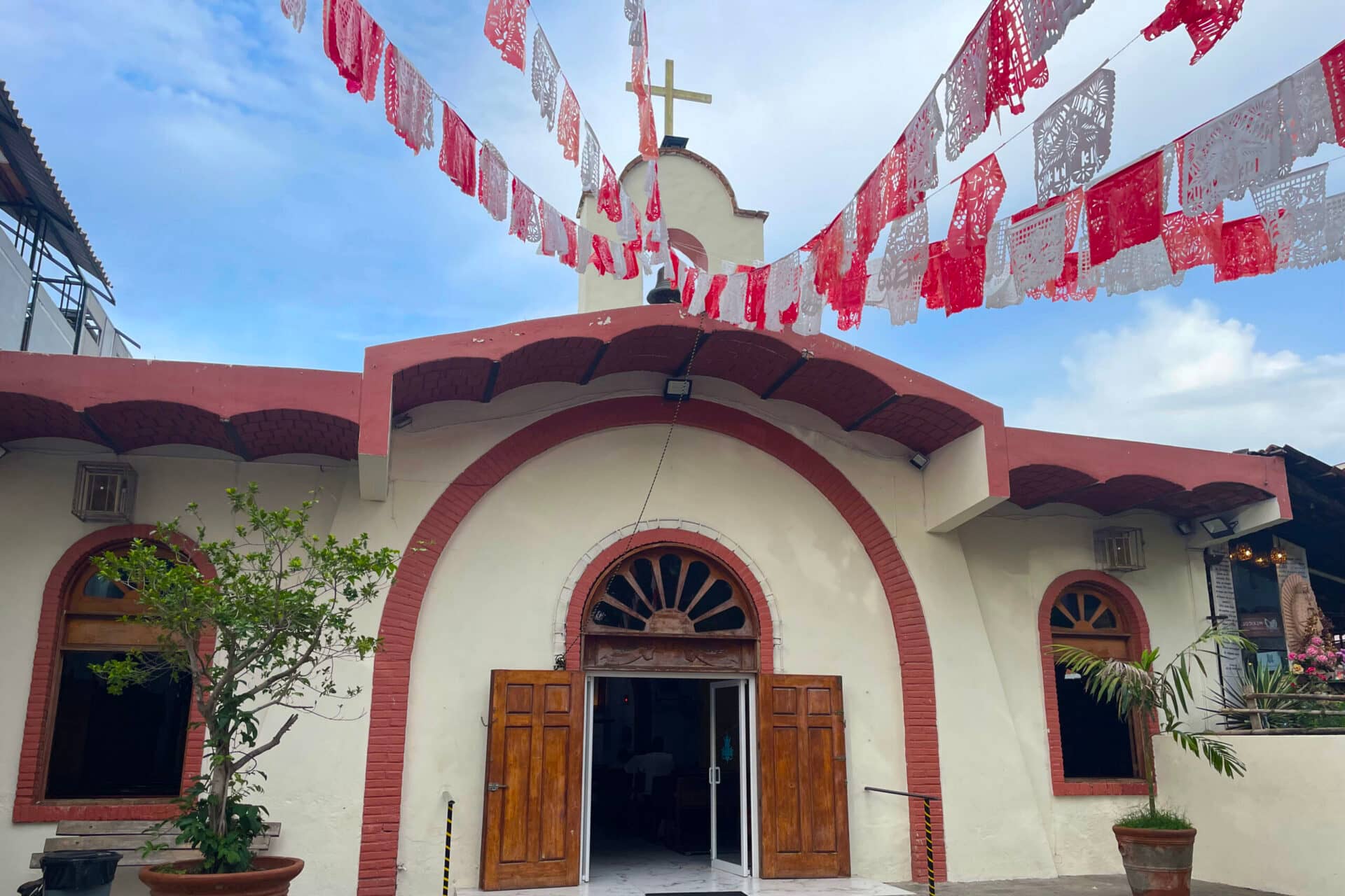 A small cream-colored church with red trim, wooden double doors, and arched windows sits under a partly cloudy sky—red and white papel picado banners flutter above, inviting memories of Mexico resorts and vibrant conference events.