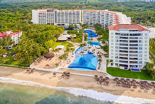 Aerial view of a beachfront resort in Mexico with two large buildings, palm trees, multiple swimming pools, sun loungers, and umbrellas on sandy beach by the ocean—perfect for meetings and conference events.