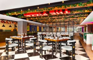 El interior de un restaurante moderno en un complejo turístico de México, con un suelo de baldosas blancas y negras, mesas de madera dispuestas para comer, sillas acolchadas, plantas verdes colgando del techo e iluminación brillante con un letrero de neón en la parte superior, perfecto para reuniones o eventos de conferencias.