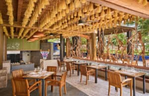 Restaurante al aire libre con mesas y sillas de madera, una singular decoración en el techo con borlas colgantes, lámparas colgantes de cobre y una pared decorativa con una colorida cabeza de toro. Ideal para reuniones o conferencias en hoteles y resorts de México.