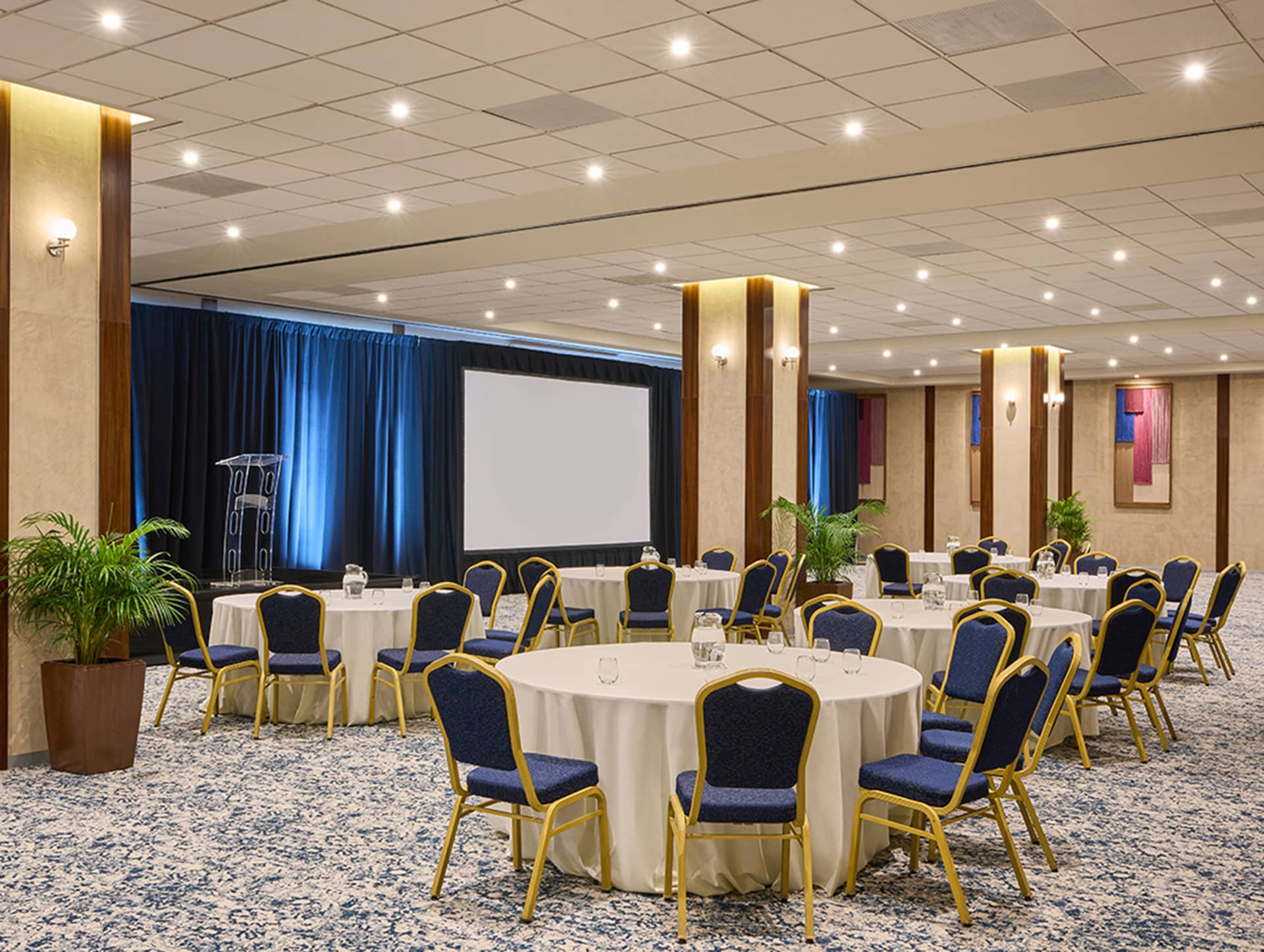 A spacious conference room at one of Mexico’s top resorts, featuring round tables and blue chairs facing a stage with a podium and large projection screen—perfect for meetings or conference events amid modern decor and potted plants.