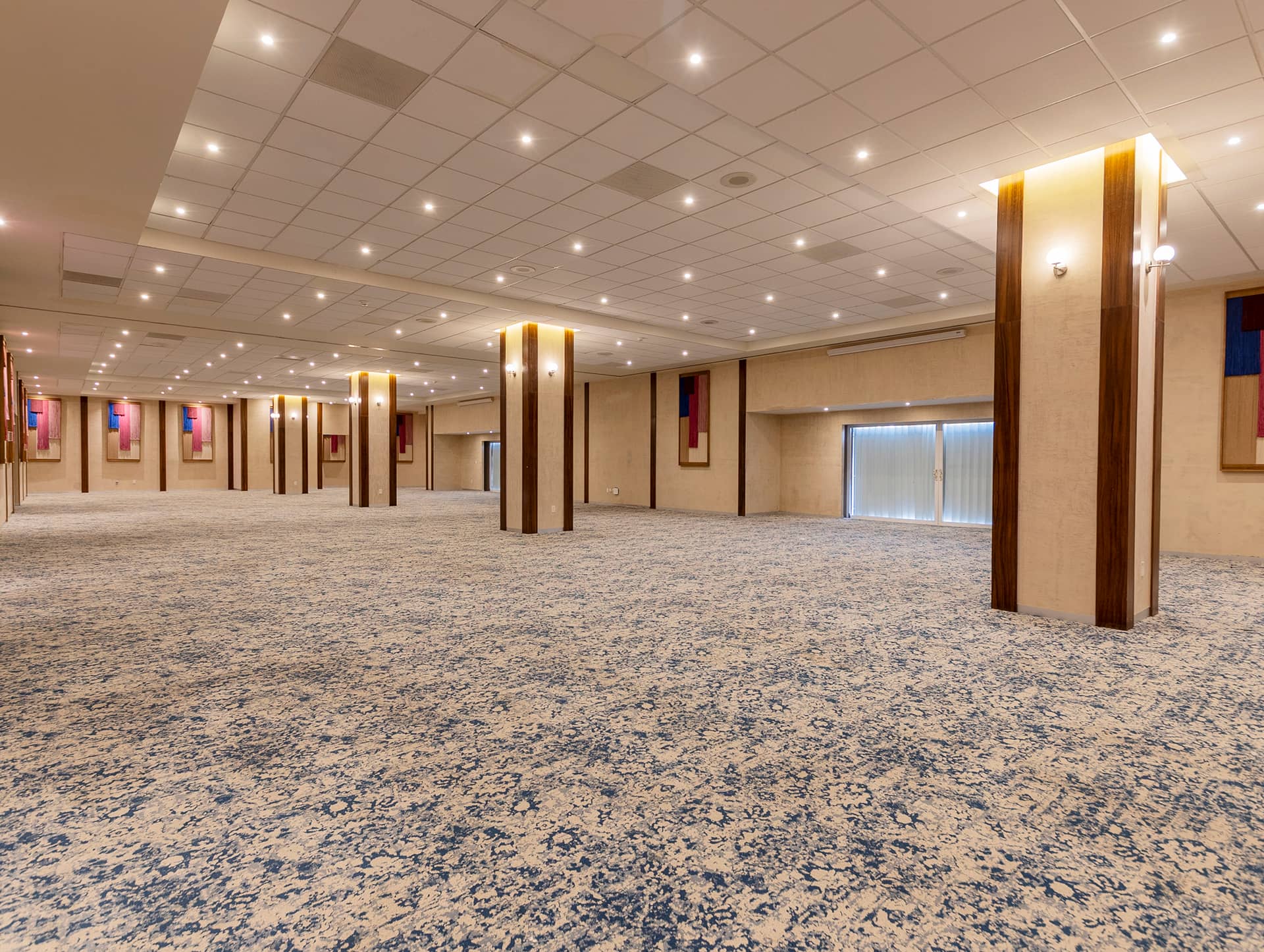 A large, empty conference room with patterned blue carpet, beige walls, and several supporting columns—ideal for meetings or conference events at Mexico hotels and resorts. Wall sconces and large windows with vertical blinds provide ample lighting.