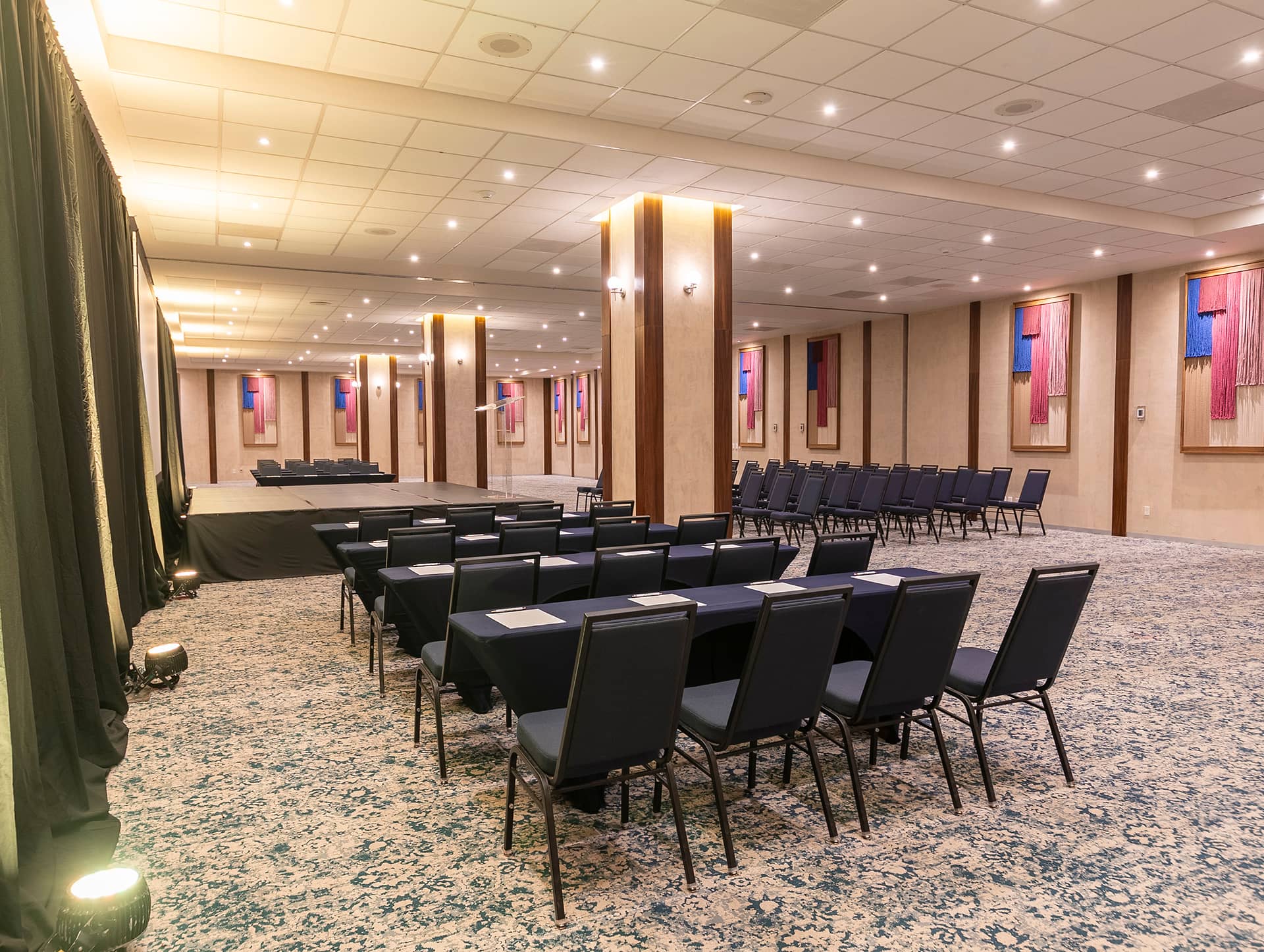 Rows of empty black chairs and tables are arranged facing a stage in a well-lit conference room at one of Mexico’s premier hotels, ideal for meetings or conference events, featuring carpeted floors, pillars, and abstract wall art.