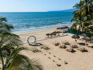 En los resorts mexicanos, la decoración para bodas en la playa incluye un arco floral, filas de sillas, mesas redondas y sombrillas de paja sobre la arena, con vistas al océano, palmeras y cielo azul; perfecta para eventos especiales o reuniones íntimas.