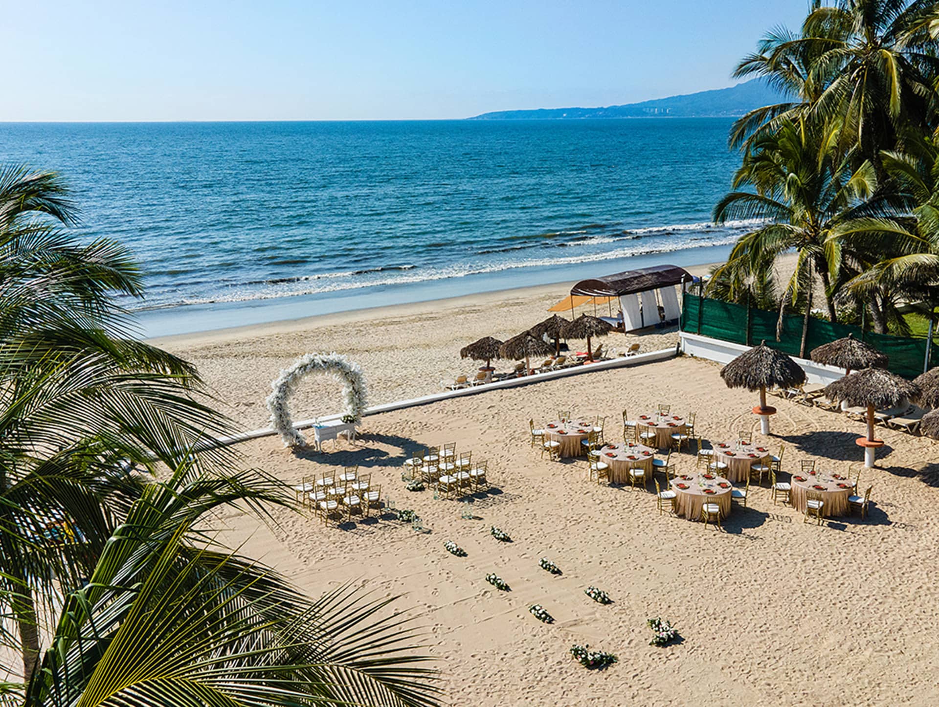 A beach wedding setup at Mexico resorts features a floral arch, rows of chairs, round tables, and thatched umbrellas on the sand, overlooking the ocean with palm trees and blue sky—perfect for special events or intimate meetings.