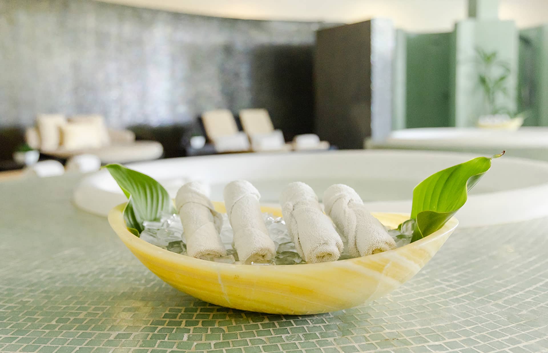 A yellow bowl filled with ice holds rolled white towels and two green leaves, set on a green mosaic counter in a spa at one of Mexico’s finest resorts, with lounge chairs and a relaxing atmosphere perfect for conference events.
