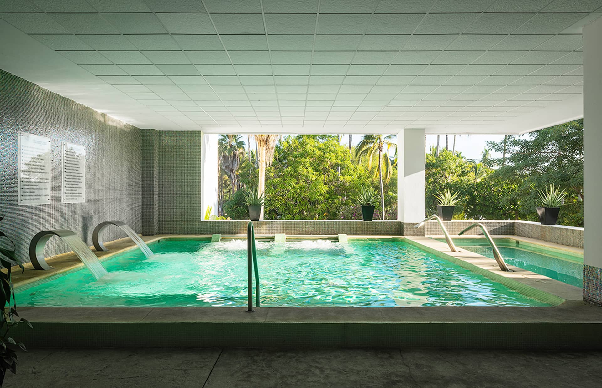 Indoor pool area with water jets and handrails, surrounded by mosaic tiles. Large windows offer views of lush green trees—perfect for relaxing between conference events at resorts or hotels in Mexico. Daylight fills the space, with potted plants along the edge.