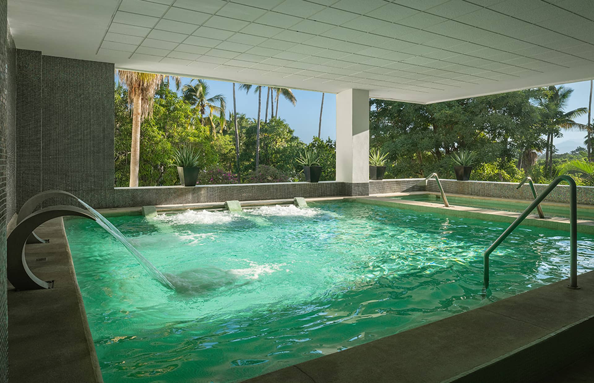 Indoor spa pool with hydrotherapy features, including water jets and curved fountains, surrounded by greenery and palm trees visible through large open windows—an ideal retreat for conference events or meetings at resorts in Mexico. Steps lead into turquoise waters.