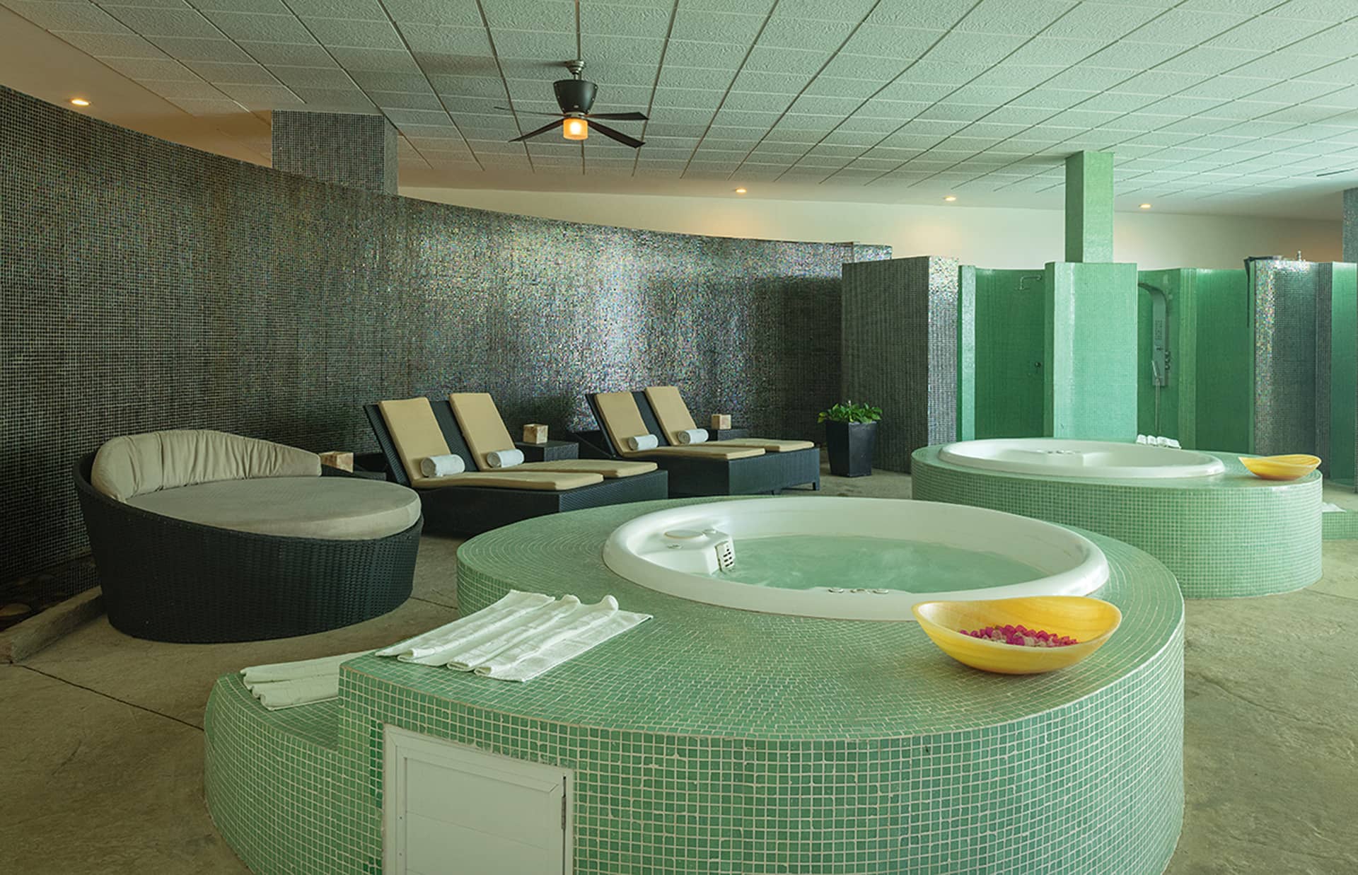A modern spa room at one of Mexico’s finest resorts, featuring two round green-tiled hot tubs, lounge chairs with rolled towels, a cushioned seat, and flower petals in the tubs—all perfect for relaxing after conference events or meetings.