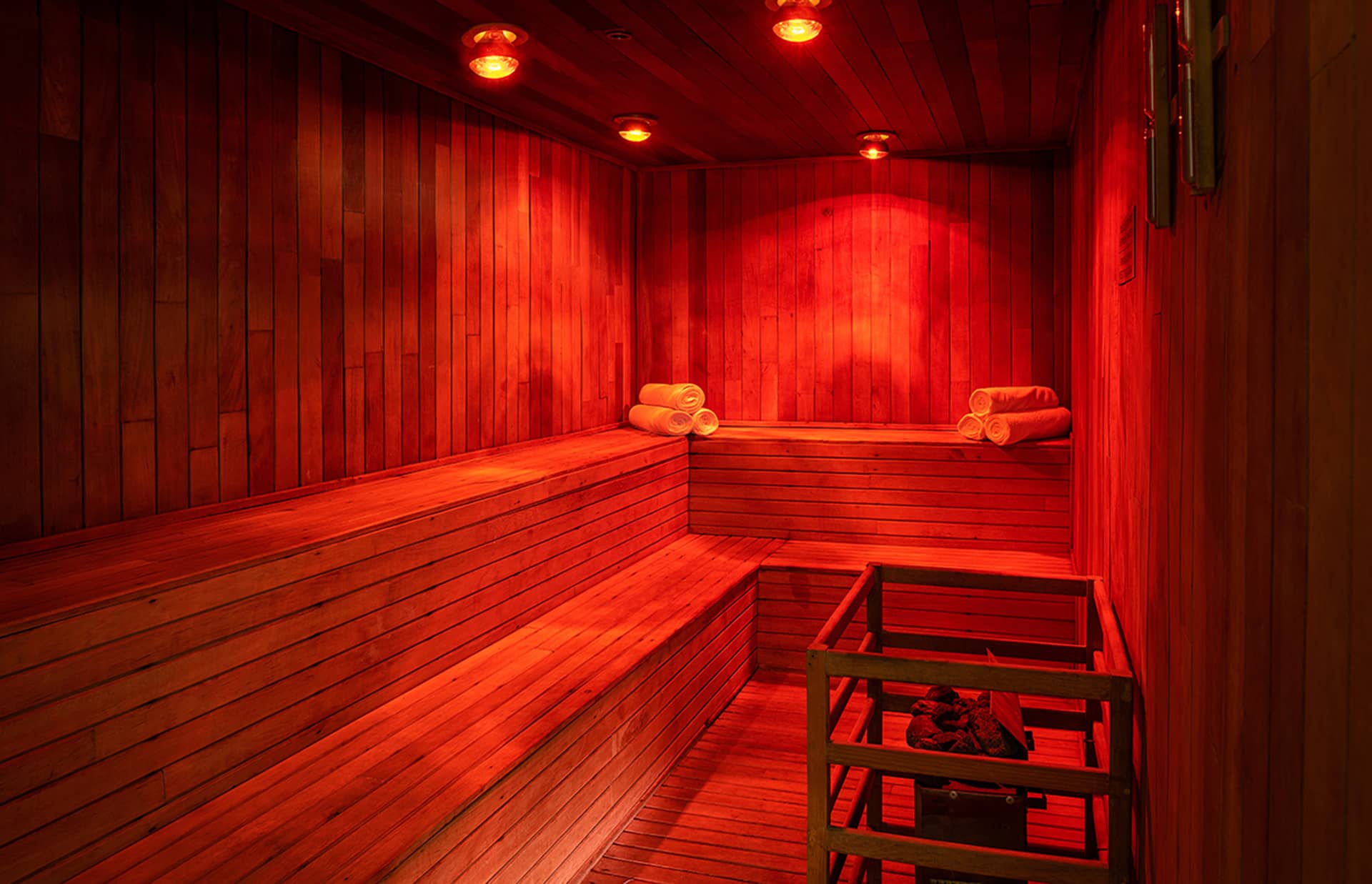 A wooden sauna room with tiered benches, neatly rolled white towels, warm red lighting, and a heater filled with stones in the corner—perfect for unwinding after conference events at resorts or hotels in Mexico.