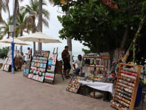 Escena de un mercado al aire libre junto al mar en un balneario mexicano, con coloridas pulseras, joyas y obras de arte en exhibición. Dos personas atienden los puestos bajo sombrillas y árboles, con palmeras al fondo y el océano visible más allá.