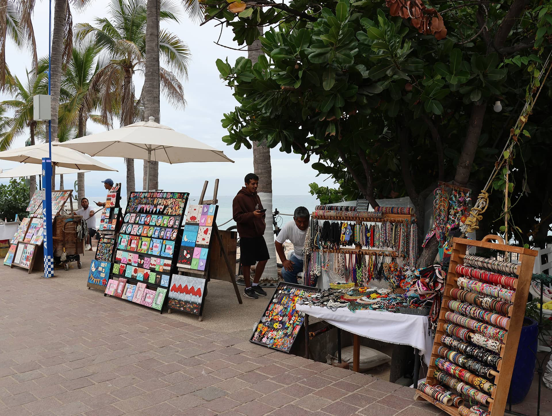Outdoor market scene by the seaside at resorts in Mexico, with colorful bracelets, jewelry, and artwork on display. Two people attend stalls under umbrellas and trees, palm trees behind them, and the ocean visible beyond.