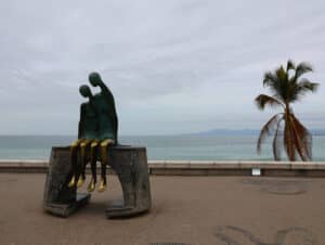 Una moderna escultura de bronce que representa dos figuras humanas abstractas se alza junto al mar, cerca de una palmera, con los hoteles y complejos turísticos mexicanos al fondo. Sus brazos y piernas dorados y alargados cuelgan sobre un banco de piedra, con las montañas y el océano de fondo, bajo un cielo nublado.