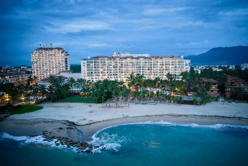 Aerial view of a beachfront resort in Mexico with palm trees, sandy beach, and gentle ocean waves, set against mountains and a cloudy sky at dusk—an ideal destination for meetings or conference events.