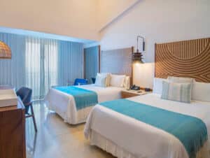 Habitación de hotel con dos camas dobles, cada una con sábanas blancas y mantas turquesas. Ideal para reuniones, la decoración moderna incluye cabeceros de madera, escritorio, sillas, lámparas de noche y una gran ventana con cortinas transparentes que dejan entrar la luz natural.